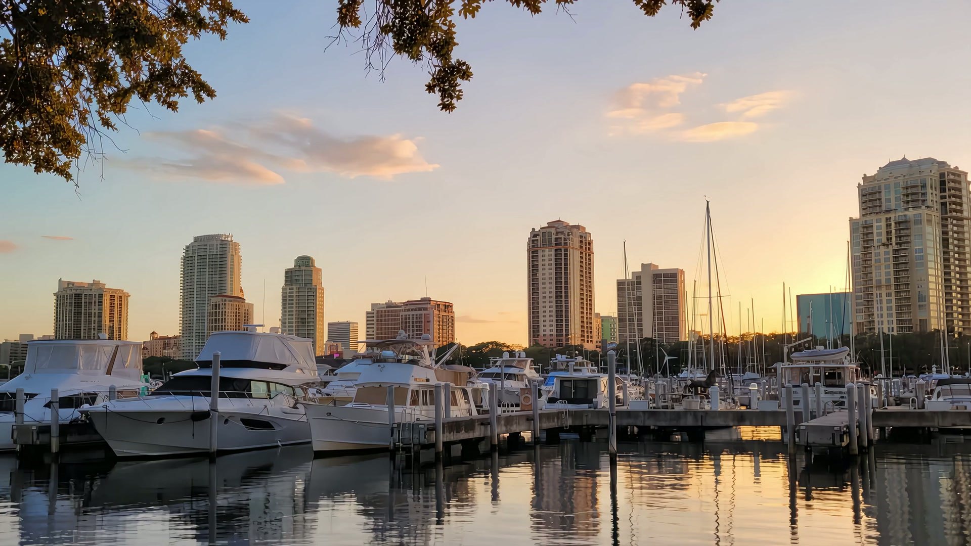A low drone view over the Vinoy Basin marina at sunset, a sailboat mast in the foreground, rising over the waterfront park toward the downtown St. Petersburg skyline